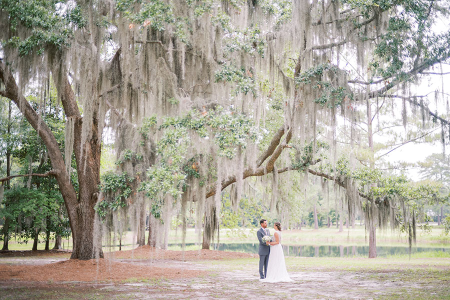 Bride and groom standing under live oaks draped with Spanish moss with a peaceful pond in the background