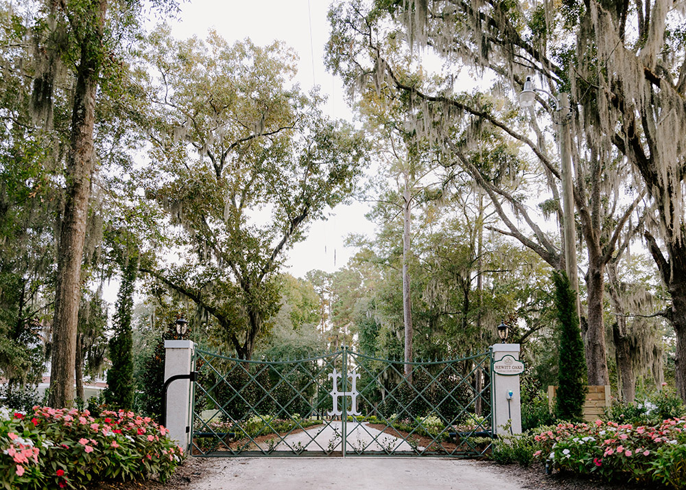 wrought iron gate at the driveway to Hewitt Oaks, surrounded by tall trees on either side and blooming pink flowers