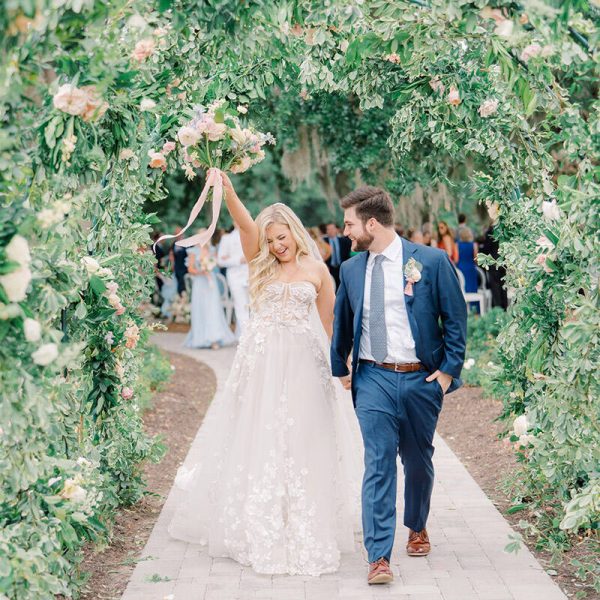 A married couple walks under a floral arch, smiling, surrounded by colorful flowers.