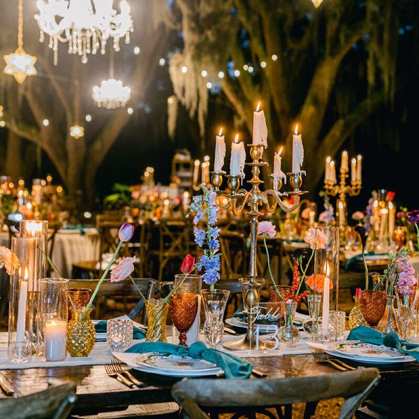 A beautifully adorned table set beneath oak trees, with hanging chandeliers casting a warm glow.