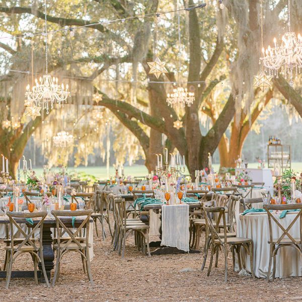 A sun-ready outdoor scene with a beautifully decorated table beneath oak trees, chandeliers suspended overhead, and the sun poised to set.