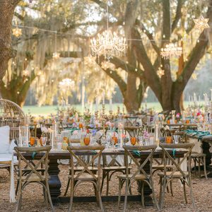Another angle of sun-ready outdoor scene with a beautifully decorated table beneath oak trees, chandeliers suspended overhead, and the sun poised to set.