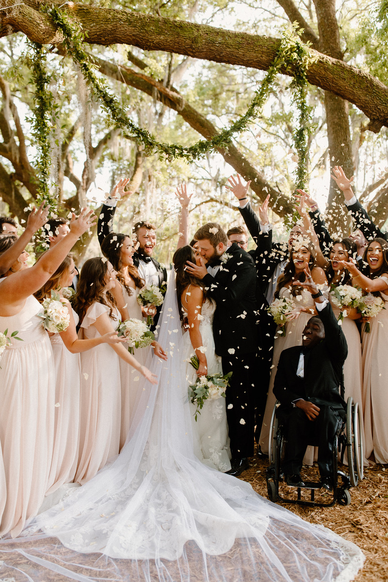 Bride and groom kissing with their wedding party cheering and confetti falling