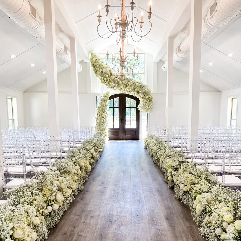Huling Chapel at Hewitt Oaks decorated for a wedding with white florals lining the aisle and a floral swoosh at the altar